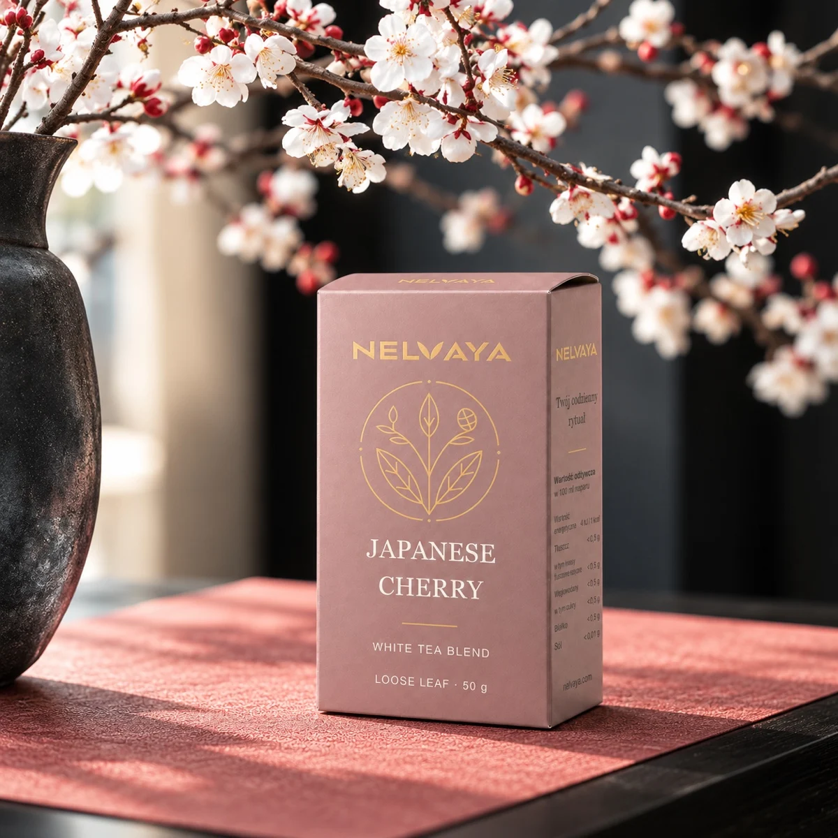 Różowy kartonik białej herbaty NELVAYA Japanese Cherry obok ciemnego wazonu z kwitnącymi gałęziami.